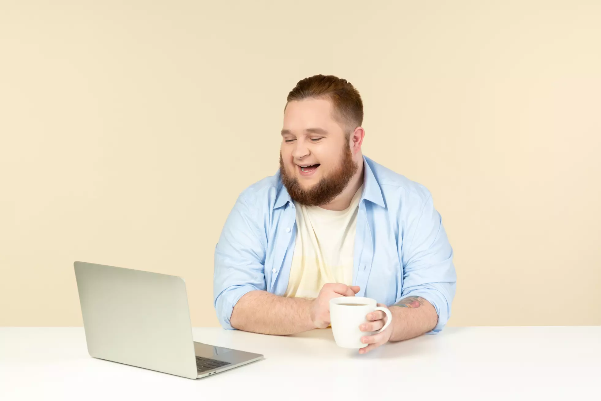 Photo of a laughing young overweight man looking at laptop and having tea